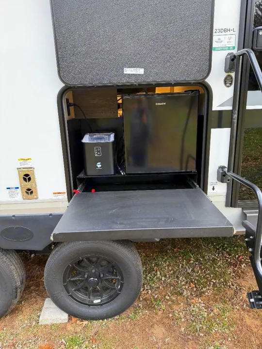This RV rental outdoor kitchen features a Hotpoint mini-fridge, ice maker, and extended pull-out shelf, visible above the wheel.. Forest River Cherokee Alpha Wolf 2025