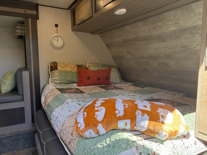 Cozy RV rental bedroom featuring a patchwork quilt, plush orange pillow with campers, and a wall clock.. Crossroads RV Sunset Trail Super Lite 2021