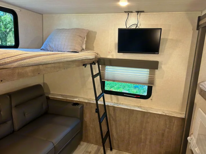 An RV rental interior shows a bunk bed with ladder, sofa below, wall-mounted TV, and window. Compact living for comfortable travel.. Forest River Surveyor 2023