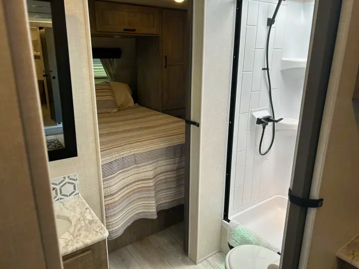 Inside an RV rental: a compact bedroom with striped bedding, adjacent to a bright shower and small vanity with a sink.. Forest River Surveyor 2023
