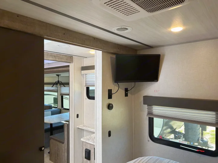 Explore this modern RV rental interior: bedroom with TV, window & exit sign, leading to a dining area. Ceiling has AC vents & lights.. Forest River Surveyor 2023