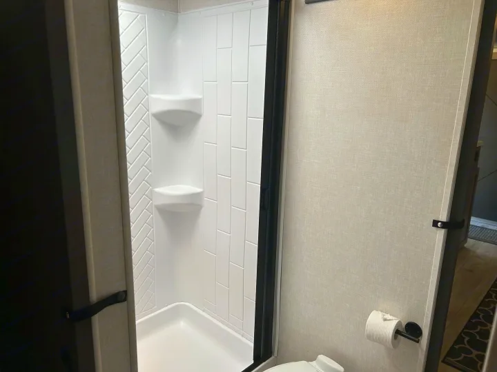 Clean RV rental bathroom features a white shower with tile-patterned walls, corner shelves, and a toilet area.. Forest River Surveyor 2023