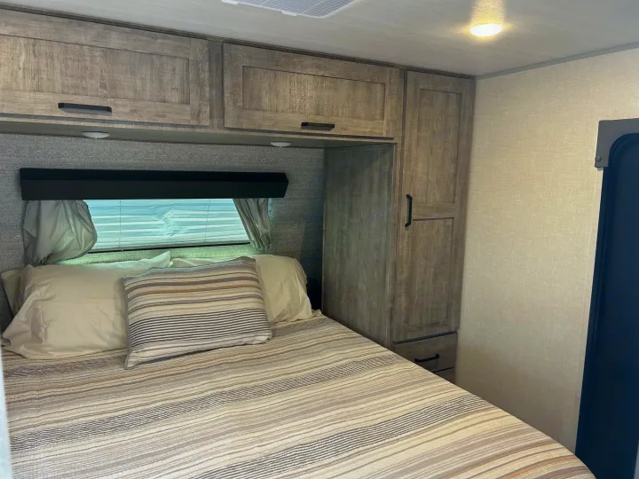 Cozy RV rental bedroom featuring a striped bed, overhead and side cabinets, and a window with blinds for comfortable travel.. Forest River surveyor Legand 2023
