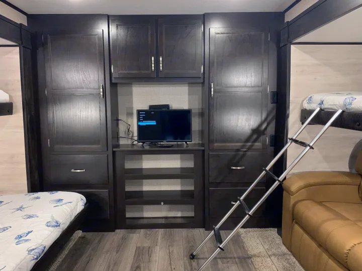 This RV rental features a cozy bunkhouse with dark wood cabinets, a TV, and ladder to upper bunk beds. Perfect for family trips.. Jayco Jay Feather 2020
