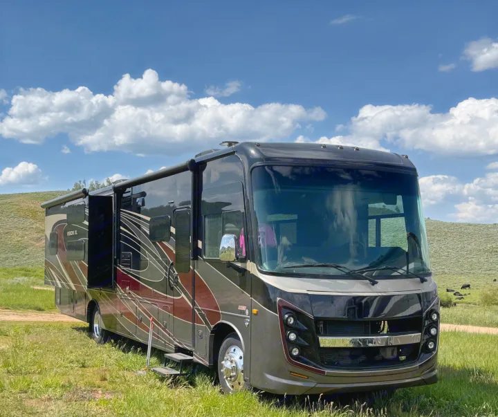 A sleek, dark RV rental with slides extended is parked in a green, hilly landscape under a bright blue, cloudy sky. Perfect for travel.. Entegra Coach Vision XL 2021