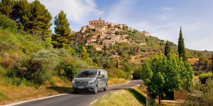 An RV rental van drives on a winding road towards a picturesque hill-top village in a sunny, green European landscape.. BAVARIA MC545BC 2026
