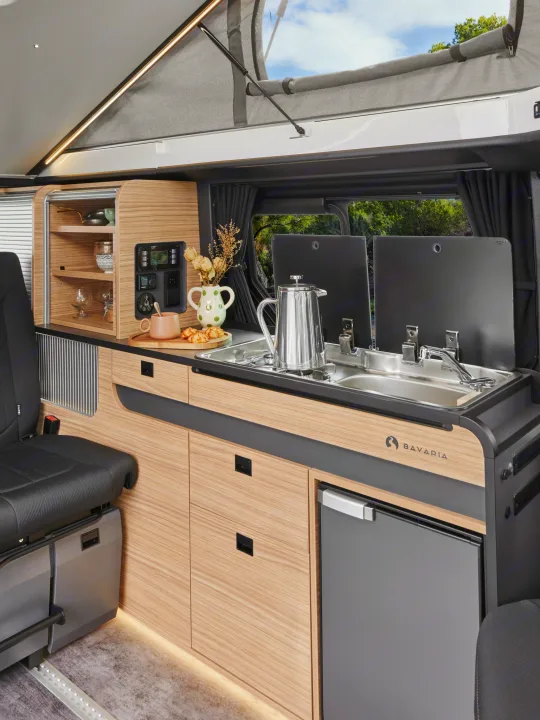 Modern rv rental interior: A compact kitchen with a sink, stove, fridge, and wooden cabinets, plus a pop-top bed above.. BAVARIA MC545BC 2026