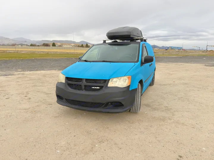 A bright blue Ram cargo van with a roof box, an ideal rv rental. Parked on a dirt lot, mountains and homes in the background.. Ram C/V Tradesman 2014