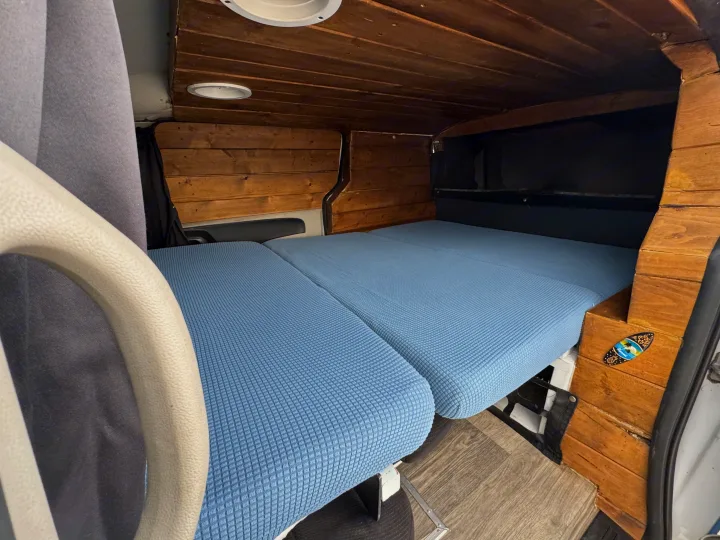 Compact rv rental interior with rustic wood paneling, featuring a blue modular bed and ceiling lights. Front handle visible.. Ram C/V Tradesman 2014
