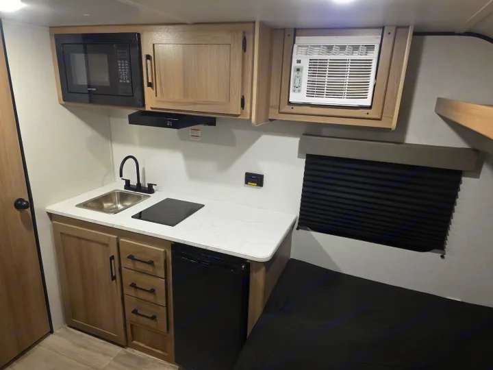Modern RV rental interior: compact kitchen w/ microwave, cooktop, sink, fridge; plus AC unit & sleeping area w/ window.. KEYSTONE COLEMAN 13B 2026