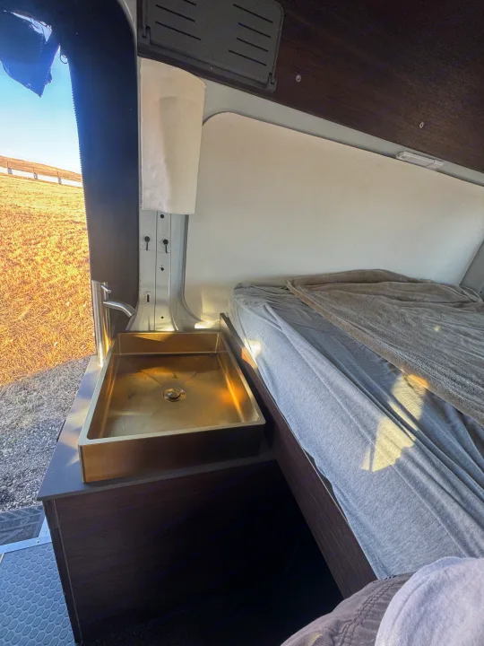 RV rental: Modern camper van interior with a golden sink, adjacent comfy bed, and bright outdoor views.. Ford Transit 2018