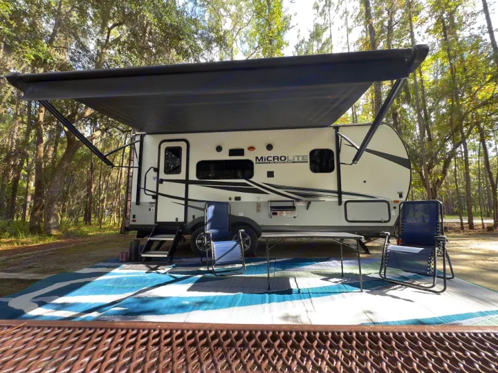 An RV rental provides a cozy campsite: a white travel trailer with an extended awning, blue chairs, and table on a rug.. Forest River Flagstaff Micro Lite 2022