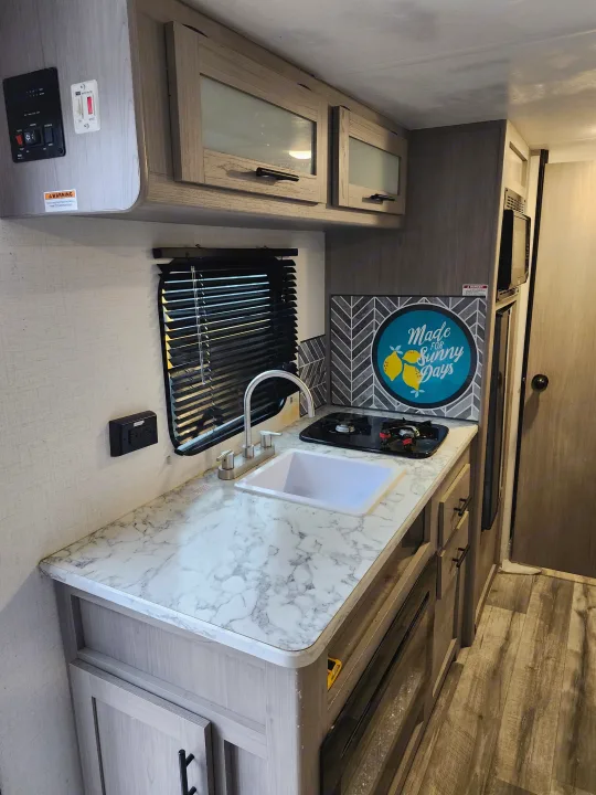 Compact RV rental kitchen: white marble-look counter, sink, stove, and light wood cabinets with microwave. Ready for your next adventure!. Dutchmen Coleman 2024