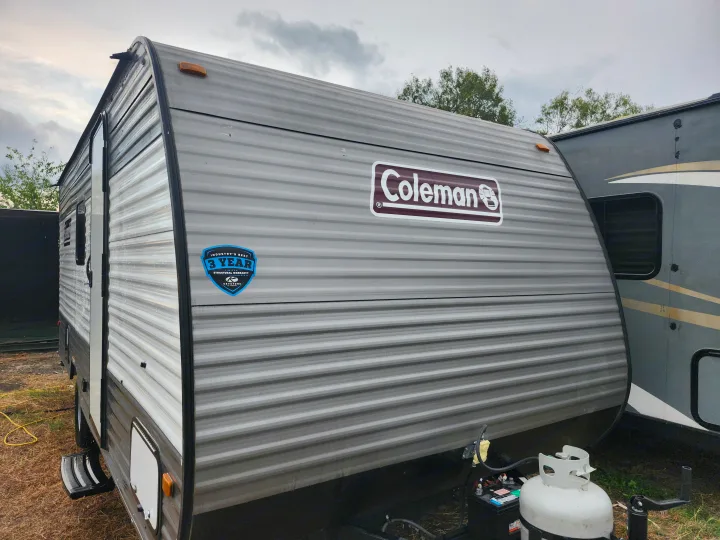 A gray Coleman travel trailer RV rental, showing a 3-year warranty badge, propane tank, and battery on the front. Parked outdoors.. Dutchmen Coleman 2024