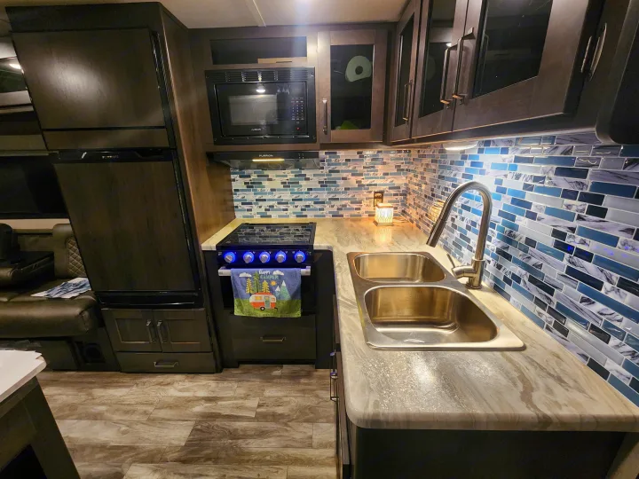 Explore this well-appointed rv rental kitchen, showcasing dark cabinets, a vibrant blue backsplash, stovetop, and double sink.. Grand Design Transcend Xplor 2022