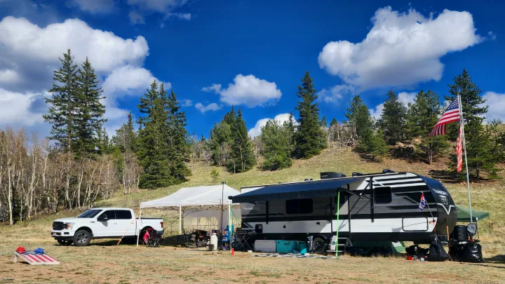 RV rental camping setup: a travel trailer, pickup truck, and canopy tent in a scenic, tree-lined clearing under a blue sky.. Grand Design Transcend Xplor 2022