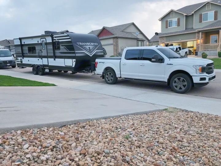 White truck towing Transcend RV on a residential street. Perfect for your next RV rental adventure!. Grand Design Transcend Xplor 2022