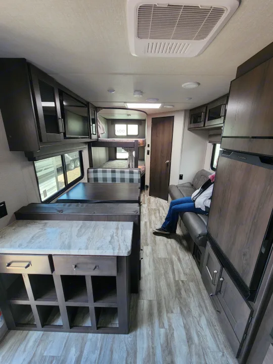 This RV rental interior features a modern dinette, bunk beds, kitchen counter, and a person relaxing on the couch.. Grand Design Transcend Xplor 2022