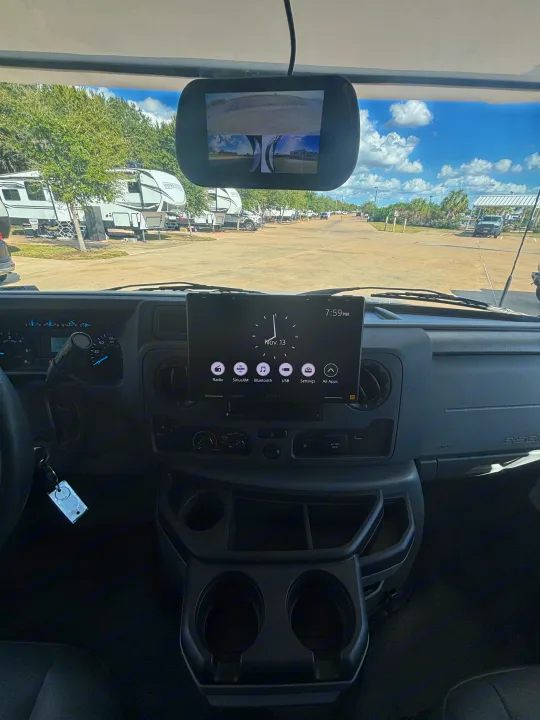 RV rental dashboard with infotainment (Nov 13, 7:59 PM), rearview monitor, and cup holders. Outside, an RV park with trailers.. Forest River Sunseeker Classic 2025