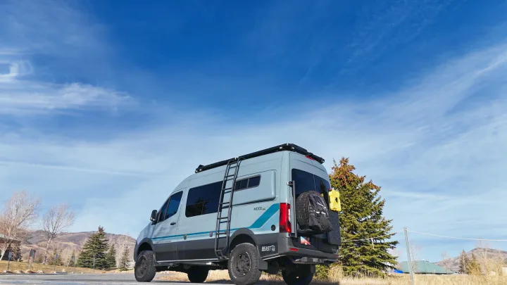 Adventure-ready gray Mode 4x4 RV rental van with off-road tires, ladder, and gear, parked under a clear blue sky.. Storyteller Overland Storyteller Overland Class B 2023