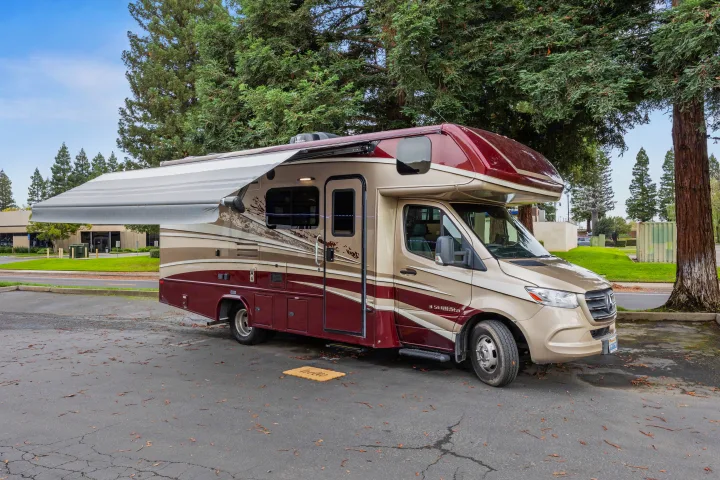 A maroon and gold Class C rv rental with an extended awning is parked outdoors on asphalt, surrounded by trees.. Dynamax Corp Isata 2021