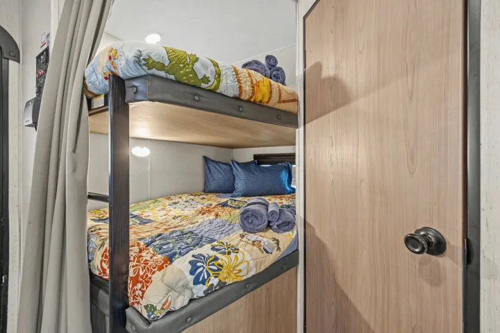 Cozy bunk beds with colorful quilts and pillows in an RV rental's bedroom area, visible next to a wooden door.. Coleman Lantern 2022