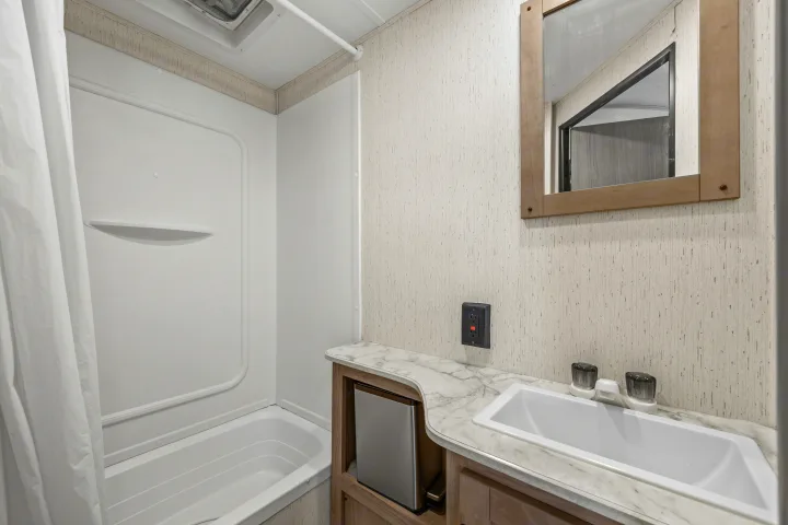 RV rental bathroom: compact shower/tub, sink vanity with mini-fridge below, mirror, and GFI outlet on textured walls.. Coleman Lantern 2022