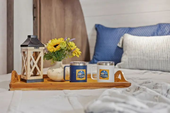 Enjoy your RV rental! A tray with "Let's Go Camping" mugs, flowers, and a lantern sits on a cozy bed.. Forest River EVO CSJT2990 2022
