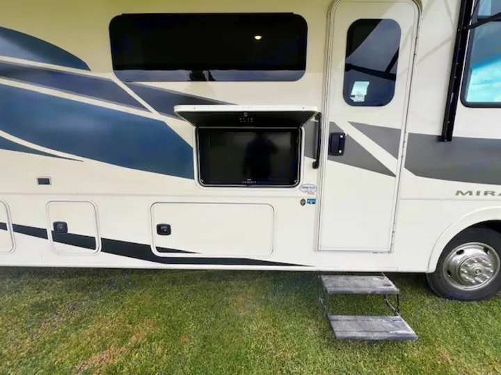 RV rental side view with an open outdoor TV, extended steps, and side storage compartments on green grass.. Forest River Coachmen Mirada 2022