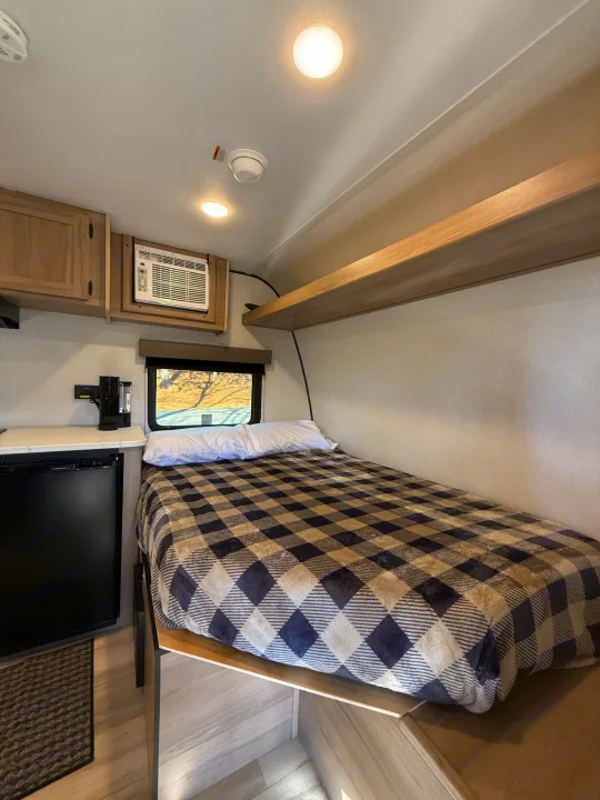Cozy RV rental bedroom featuring a checkered bed, window, AC, fridge, and overhead storage for a comfortable stay.. Keystone Coleman 13R 2026
