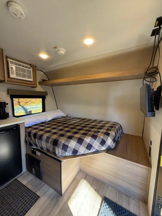 Cozy RV rental interior featuring a comfortable bed with plaid blanket, AC unit, TV, and small fridge.. Keystone Coleman 13R 2026