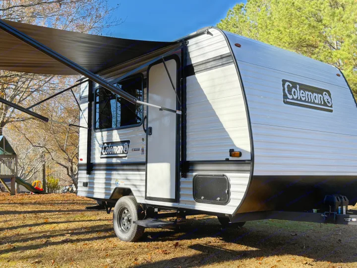 A white and grey Coleman travel trailer, ready for an RV rental adventure, parked outdoors with its awning extended.. Keystone Coleman 13R 2026