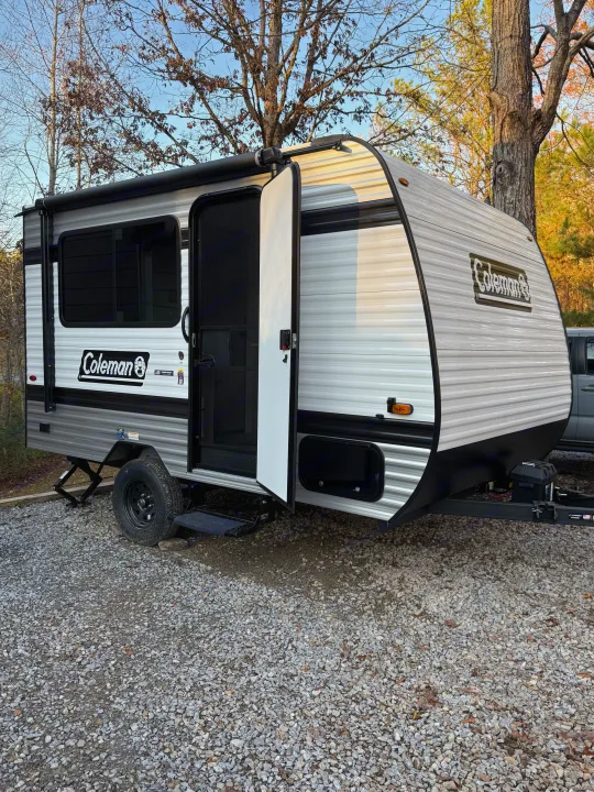 Grey and white Coleman travel trailer RV rental parked on a gravel camping site, door open, under bare trees.. Keystone Coleman 13R 2026