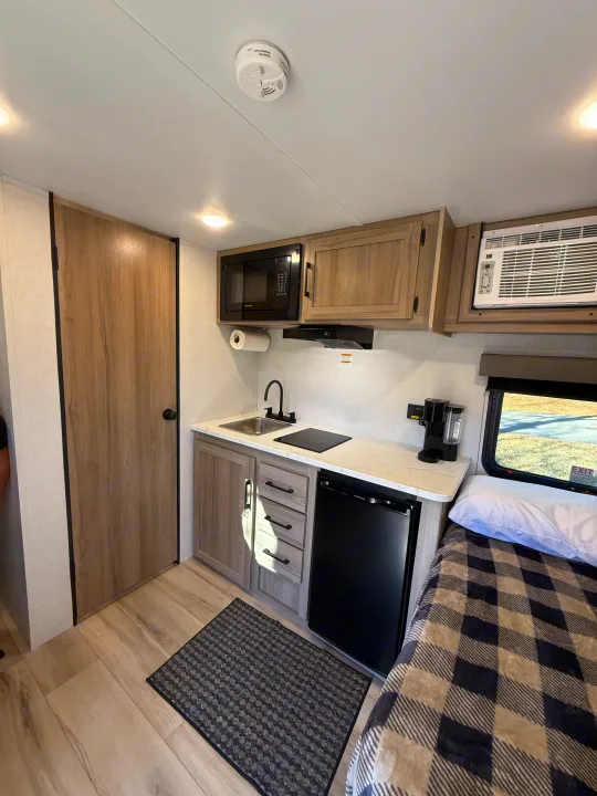 Cozy rv rental space, featuring a kitchen area with microwave, sink, mini-fridge, and a snug bed with plaid bedding.. Keystone Coleman 13R 2026