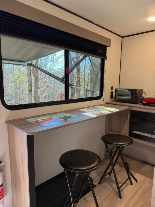 Cozy rv rental interior featuring a bar with stools, forest views, and a small kitchen setup. Perfect for glamping!. Keystone Coleman 13R 2026