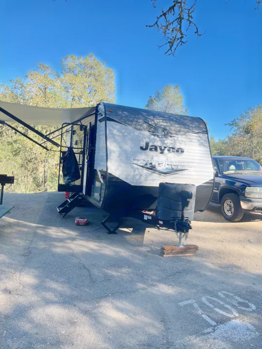 A Jayco RV rental with its awning extended is parked at campsite 708, with a truck beside it and trees around.. Jayco Jay Flight SLX Rocky Mountain Edition 2022