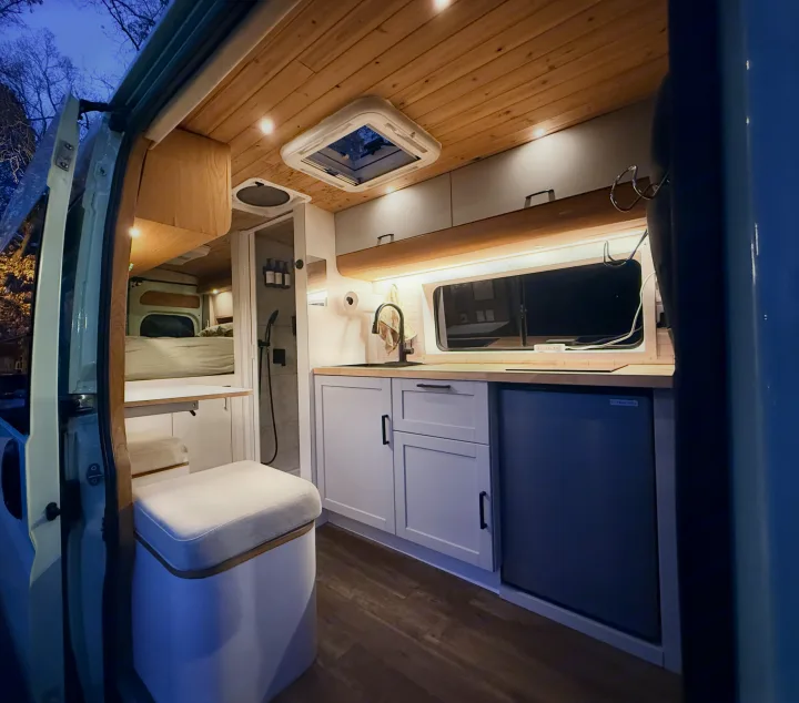 RV rental interior: A modern camper van with a compact kitchen, comfortable bed, & a shower, bathed in warm lighting.. Dodge Pro Master High Roof 3500 Extended 2021
