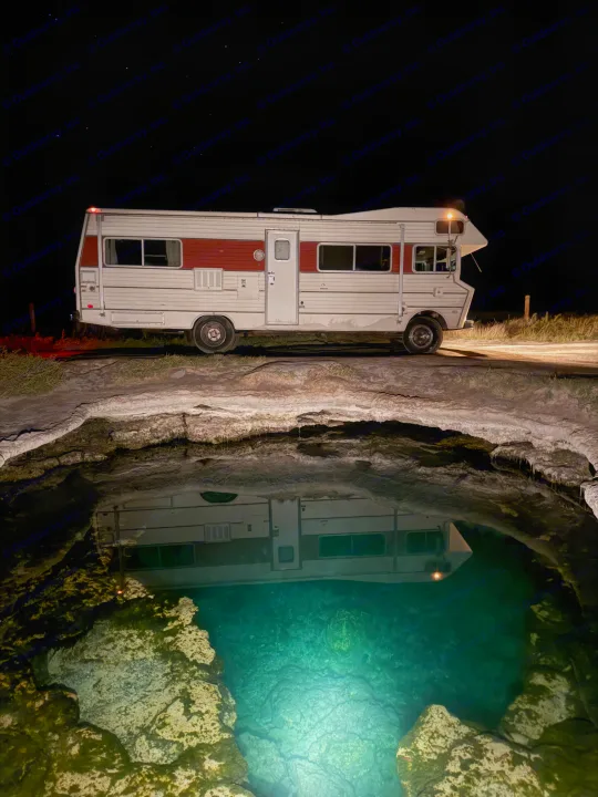 Night shot: RV rental parked by a glowing blue hot spring, reflecting on the water under a starry sky.. International 510A 1973