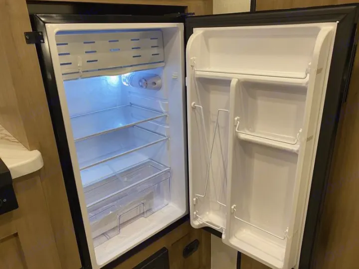 Empty and illuminated refrigerator within an RV rental, showing internal shelves, crisper drawer, and door storage for your travel needs.. Forest River Cherokee Wolf Pup 2025