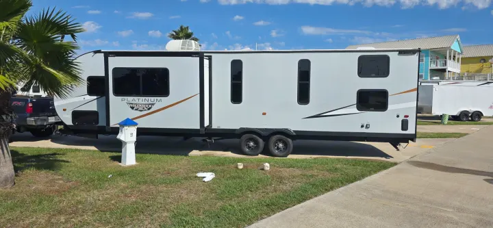 Grey Wildwood Platinum rv rental with an extended slide-out. Parked on concrete at a sunny RV resort, palm trees and colorful homes in view.. Forest River Wildwood FSX Platinum 2024