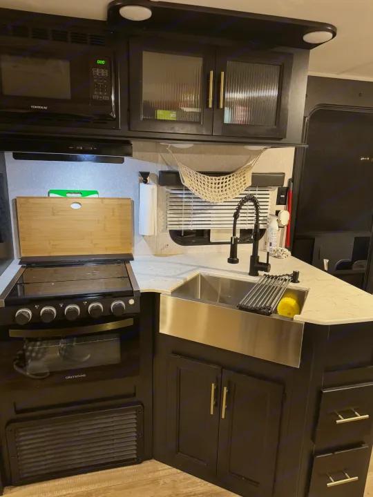 RV rental kitchen: black microwave, stove, stainless steel farmhouse sink, and dark wood cabinets.. Forest River Cherokee Grey Wolf 2022