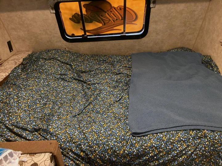 Cozy RV rental bed, featuring a dark floral patterned comforter and a folded grey knitted blanket, ready for your trip.. Clipper 17BH 2018