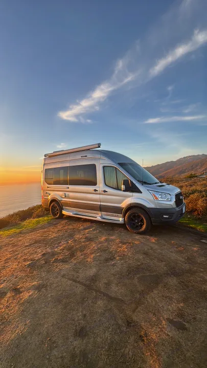 Silver RV rental van on a cliff, ocean sunset & hills in background. Ideal for scenic road trips & adventure.. Pleasure-Way Pleasure-Way Class B 2023