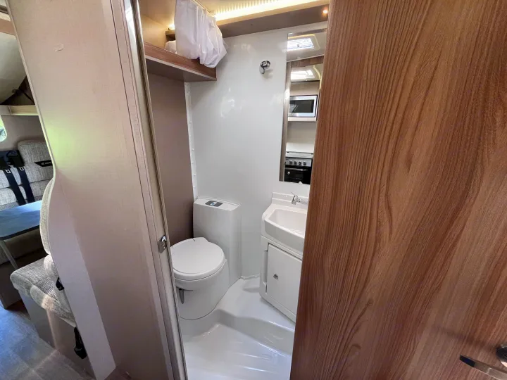 Inside this RV rental, a compact white bathroom features a toilet and sink. A mirrored wall reveals the kitchen with microwave and stove.. Swift Edge 486 2024