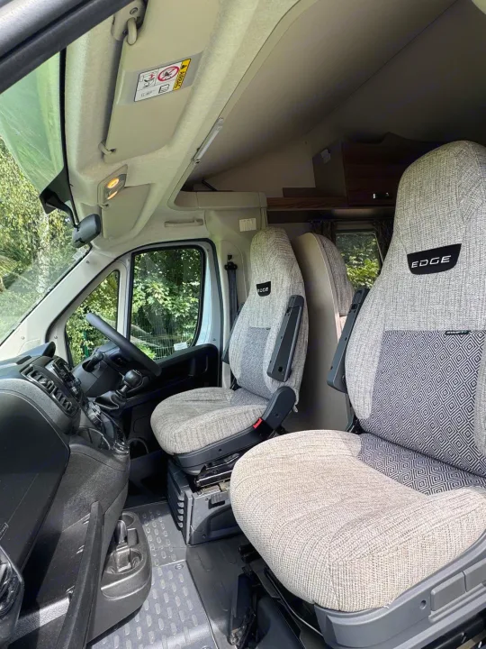 The front cab of an RV rental, featuring light-colored driver and passenger seats with "EDGE" branding, dashboard, and steering wheel.. Swift Edge 486 2024