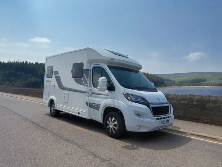 White Xplora motorhome (RV rental) parked by a scenic lake and green hills under a clear sky, ideal for outdoor adventures.. Moto-Trek Xplora FDB 2025