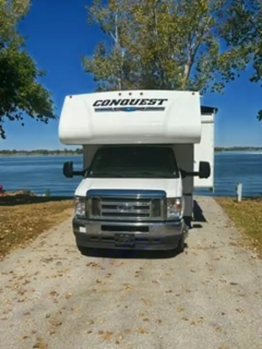 A white Conquest Class C **rv rental** is parked on a leafy path by a beautiful lake, ready for your next adventure.. Gulf Stream Conquest 2022