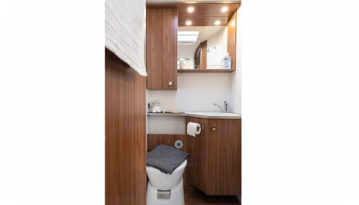Inside an RV rental: a compact bathroom with a white toilet, corner sink, mirrored cabinet, and warm wooden storage.. PILOTE P726 C 2026