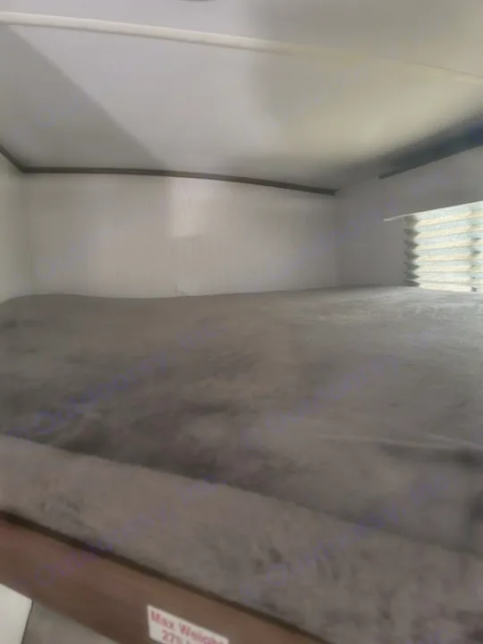 Empty bunk bed in an RV rental, featuring a dark grey blanket, white walls, and a window blind. Max weight 275 lbs.. Cruiser RV MPG 2023