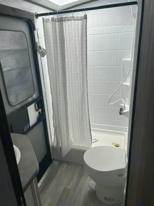 This RV rental bathroom features a white toilet, shower with a striped curtain, and a small sink, offering compact convenience.. Cruiser RV MPG 2023